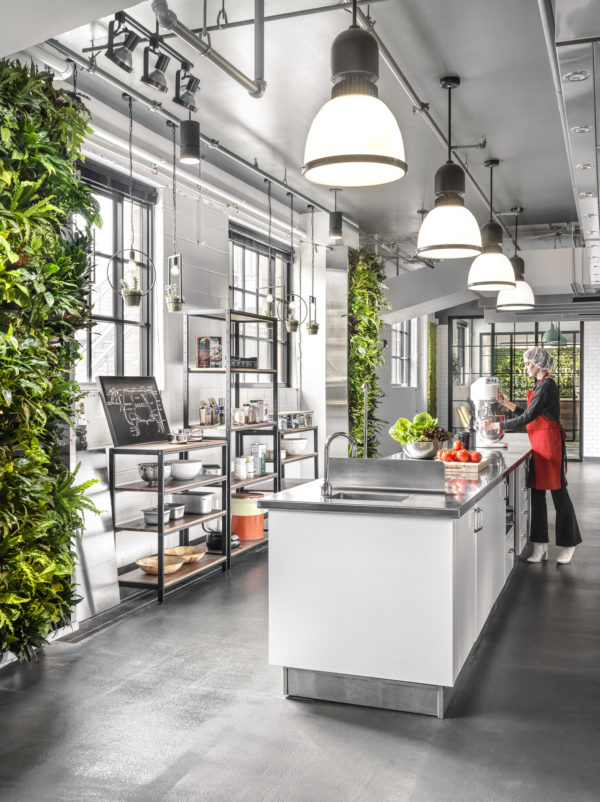 A woman prepares food in a modern test kitchen, with accents of plant life extending from the wall contrasting the sleek decor of the kitchen